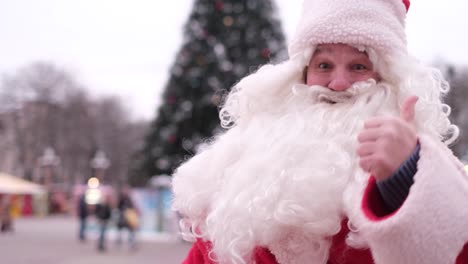 stylish santa claus with a white beard in a red suit showing thumb up merry christmas 2023 stylish santa claus with a white beard in a red suit showing thumb up on the background of the city christmas tree merry christmas 2023