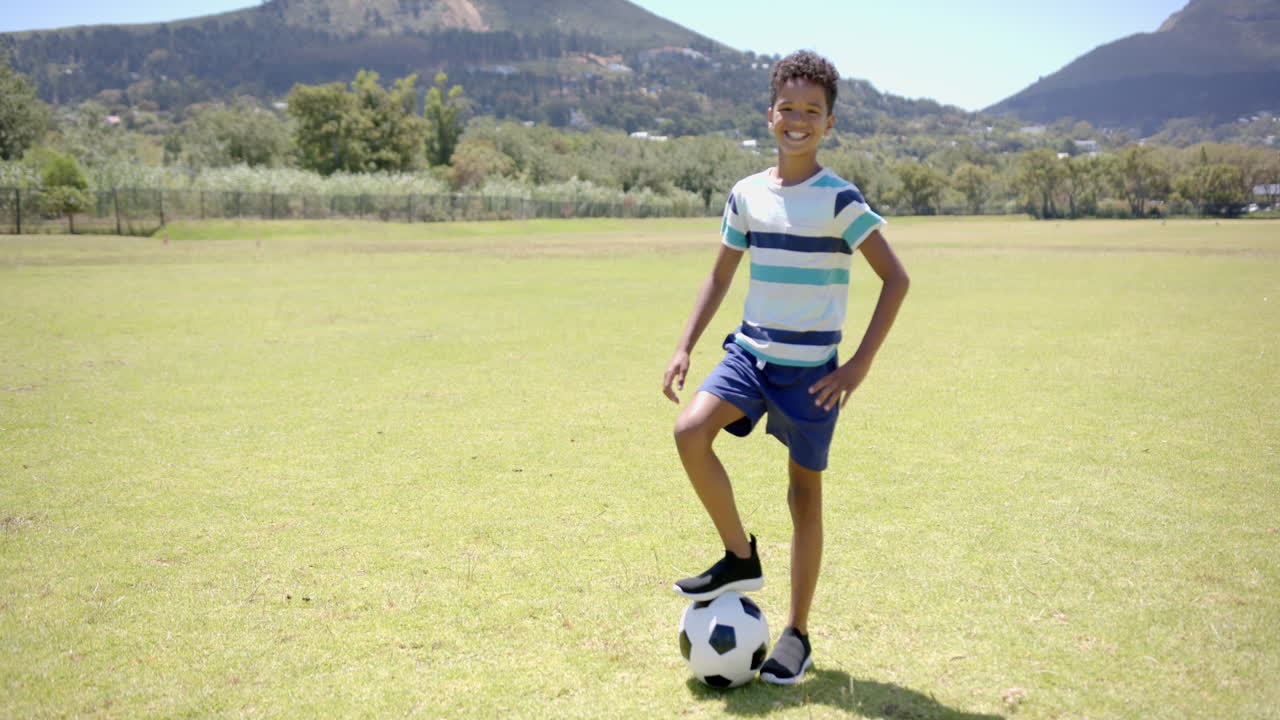 Biracial boy with curly hair smiles, soccer ball at foot, in sunny school park