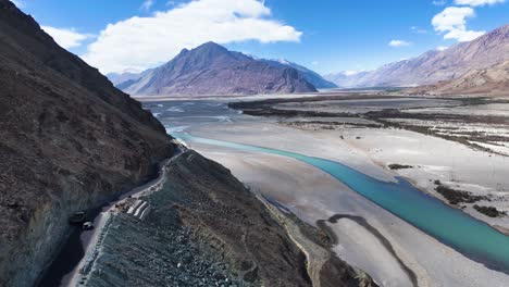 aerial drone shot of a car driving near the crystal-clear river in nubra valley, surrounded by mountains and sand dunes.