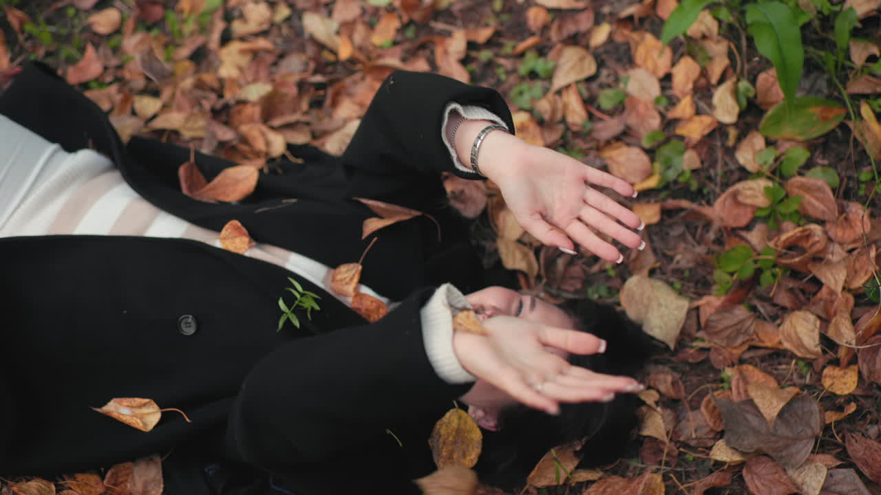 Casual lady lies on forest ground smiling, hands raised to block falling dry leaves, relaxed mood among autumn foliage, cozy coat and sweater, playful moment captured from above with soft greenery