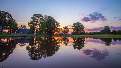 Ein-Leuchtend-Orangefarbener-Sonnenaufgang-Hinter-Den-Bäumen-Am-Ufer-Eines-Sees-Mit-Einem-Haus