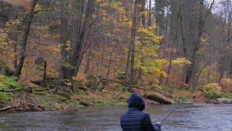 A Middleaged Man in a Black Jacket Throws a Fishing Rod Into the River on a Gloomy Autumn Day. A middle-aged man in a black jacket throws a fishing rod into the river on a gloomy autumn day. Male fishing as a hobby. Fishing on the river in the middle of a picturesque forest