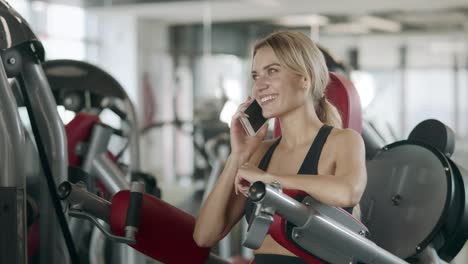 Fröhliche-Frau,-Die-Am-Fitness-Trainingsgerät-In-Der-Sporthalle-Mit-Dem-Mobiltelefon-Spricht.