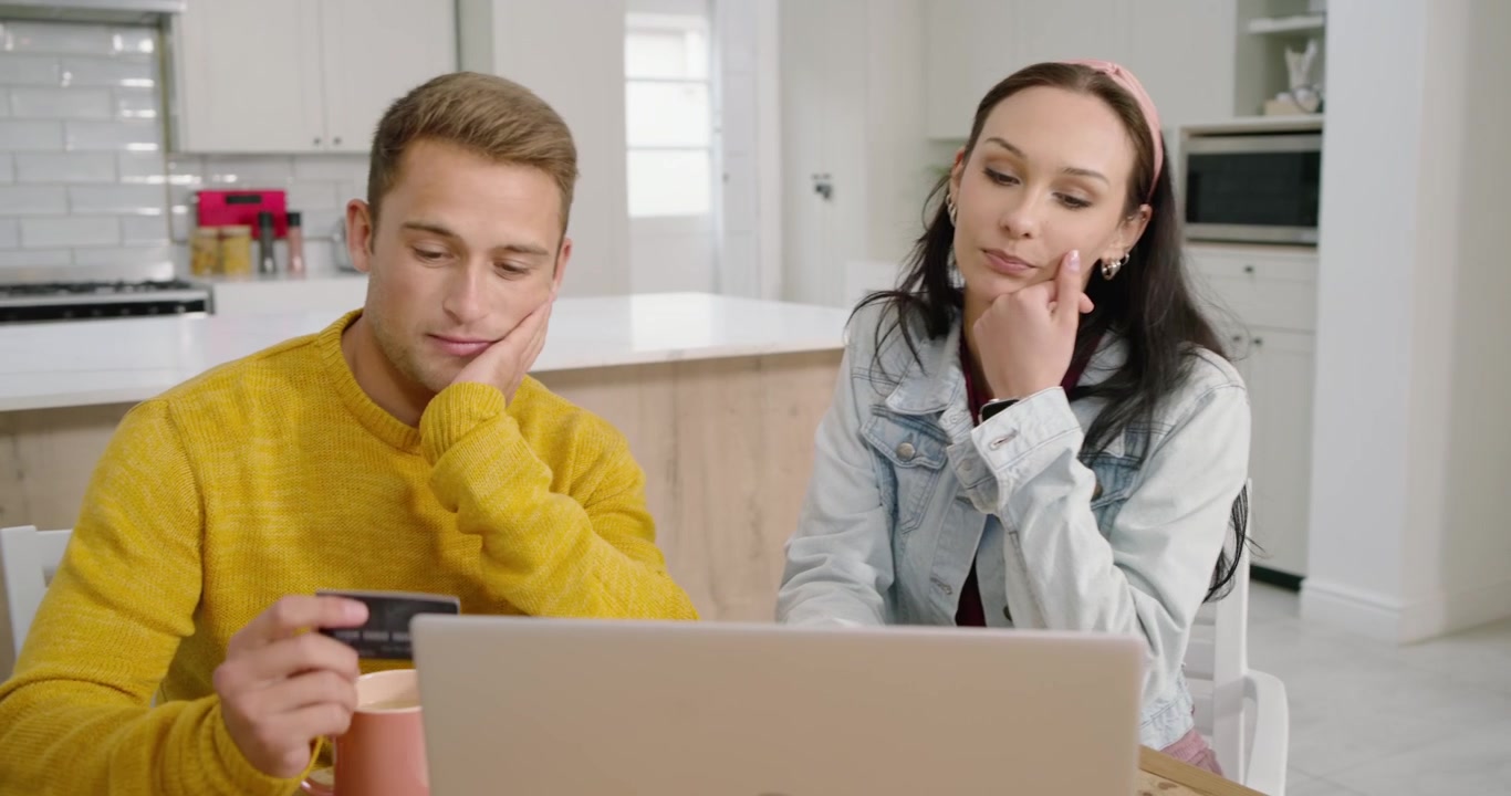 Coppia a un tavolo da cucina in legno, l'uomo con un maglione color senape mostra una carta e digita sul laptop per pagare.