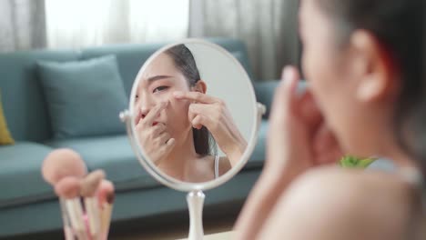 거울에 반사 된 걱정 된 젊은 아시아 여성은 집에서 메이크업을 할 때 여드름을 니다.