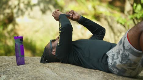 A man lays down and checks his phone on a giant rock in the middle of the woods
