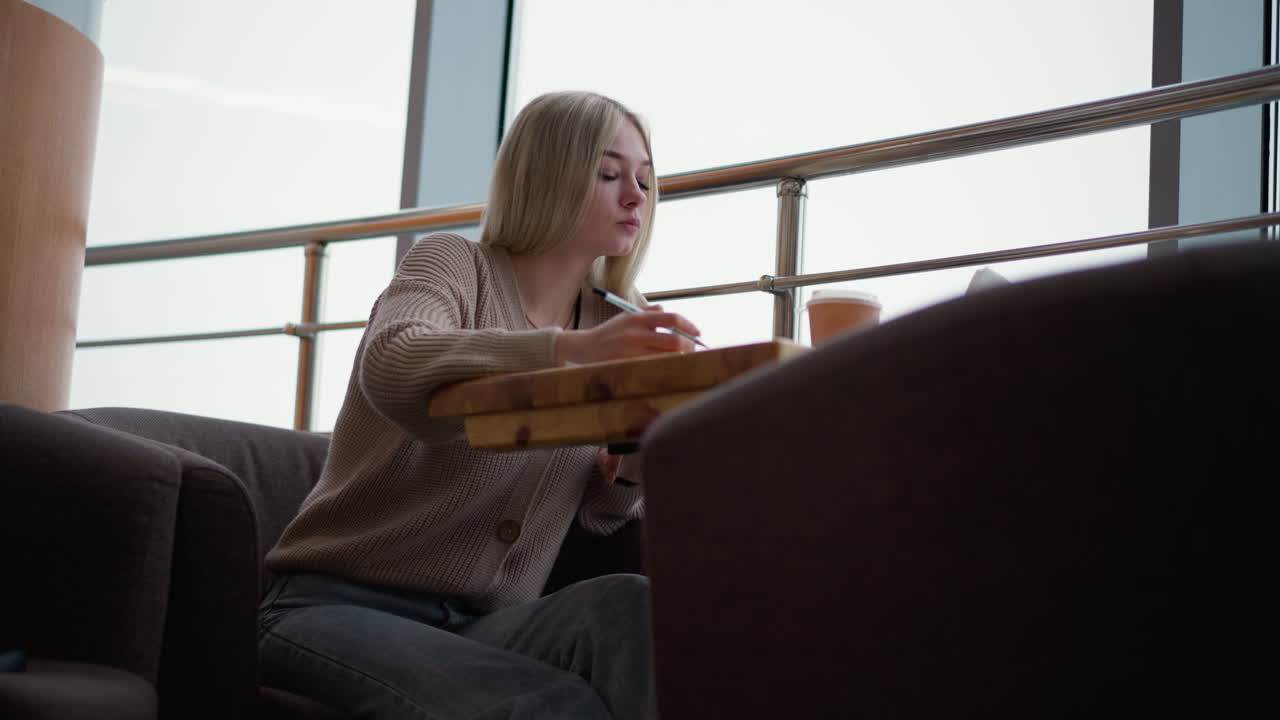 커피를 마시면서 쇼핑몰에서 노트북에 사려 깊게 쓰는 젊은 여성은 쾌적한 도시 카페 환경에서 밖의 전망을 보여주는 유리 창문으로 작업에 집중합니다.