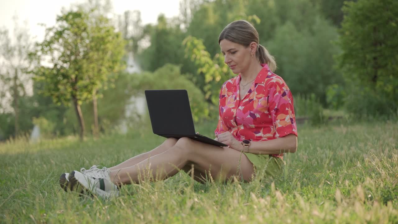 una mujer de mediana edad trabaja usando su portátil en un parque público. ella escribe y piensa.