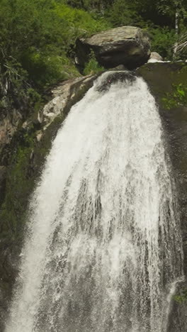 narrow waterfall on high steep rock in green summer forest slow motion. whitewater falls down from cliff ledge in nature park on sunny day. water flow