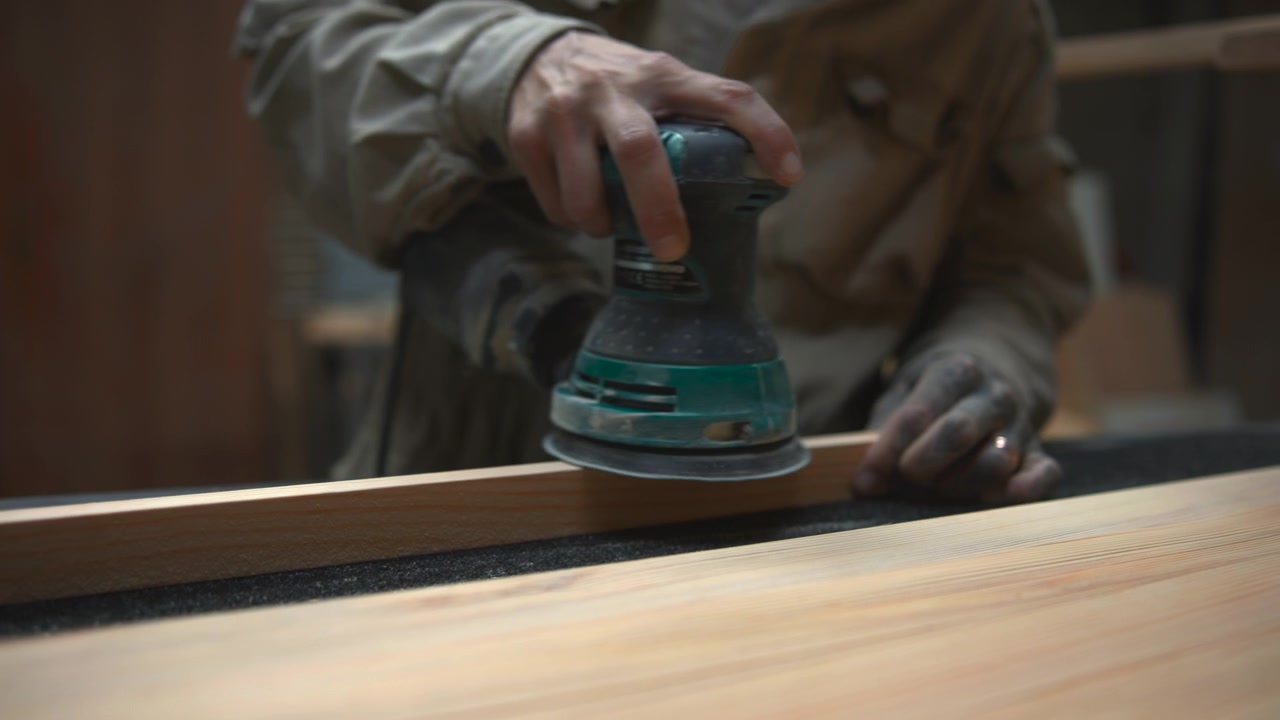 Manos sujetando tabla de madera mientras una lijadora eléctrica de palma hace contacto cercano con la superficie, creando polvo fino, desenfoque suave en el borde de la almohadilla, luz cálida de taller y trabajo de acabado de carpintería enfocado.