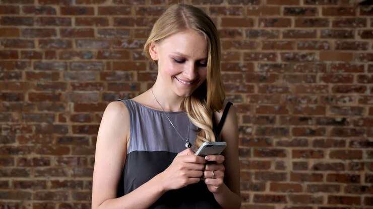 Young cute blond girl is typing message on smartphone, thinking concept, communication concept, brick background