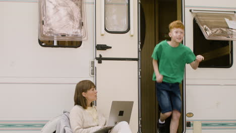 concentrated woman sitting on chair and working on laptop computer at the camping in the forest while her son getting out of the campervan