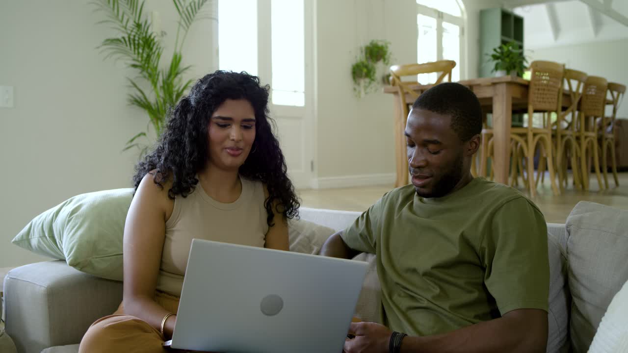 Diverse couple sitting on couch, man tapping phone, sharing screen while woman using silver laptop