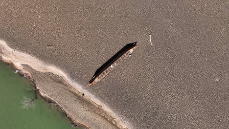 Descending-aerial-view-over-abandoned-wooden-boat-laying-on-dry-cracked-terrain-in-the-Sau-reservoir,-Spain