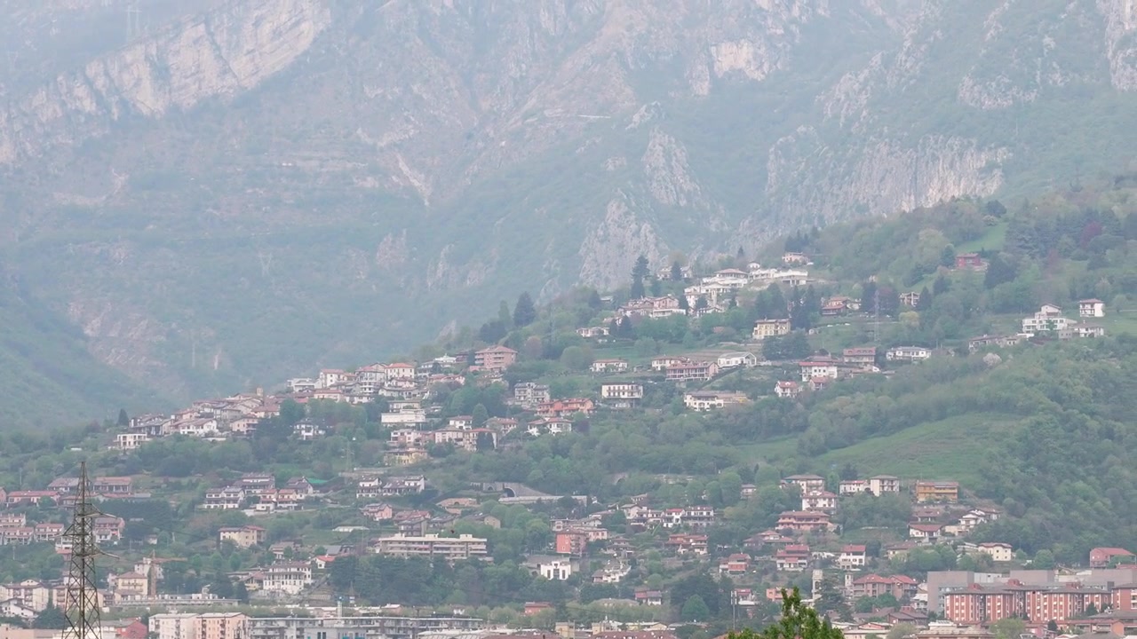 Ampie riprese grandangolari catturano una cittadina collinare punteggiata da edifici colorati e alberi rigogliosi, annidata sotto imponenti montagne nebbiose, sotto una luce diurna morbida e naturale, con una composizione stabile.