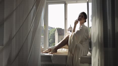 Bride-in-boudoir-dress-sitting-on-window-sill-wedding-morning-preparations-woman-in-night-gown,-veil