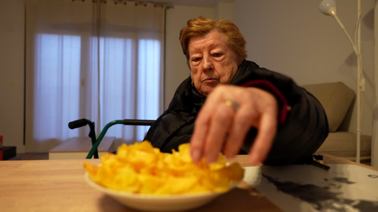 Grandmother in wheelchair taking potato chips from a bowl at home