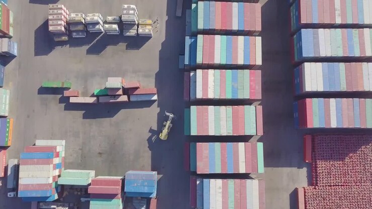 A container cargo carrier forklift machine moves through a large container yard while many truck wait to be loaded