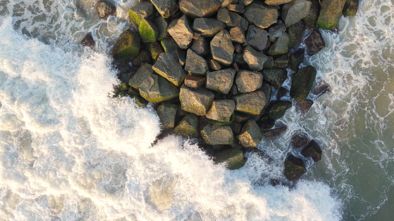 Top-down drone shot of ocean waves crashing against a rocky breakwater on the coast of Sri Lanka, featuring white sea foam and green mossy stones.