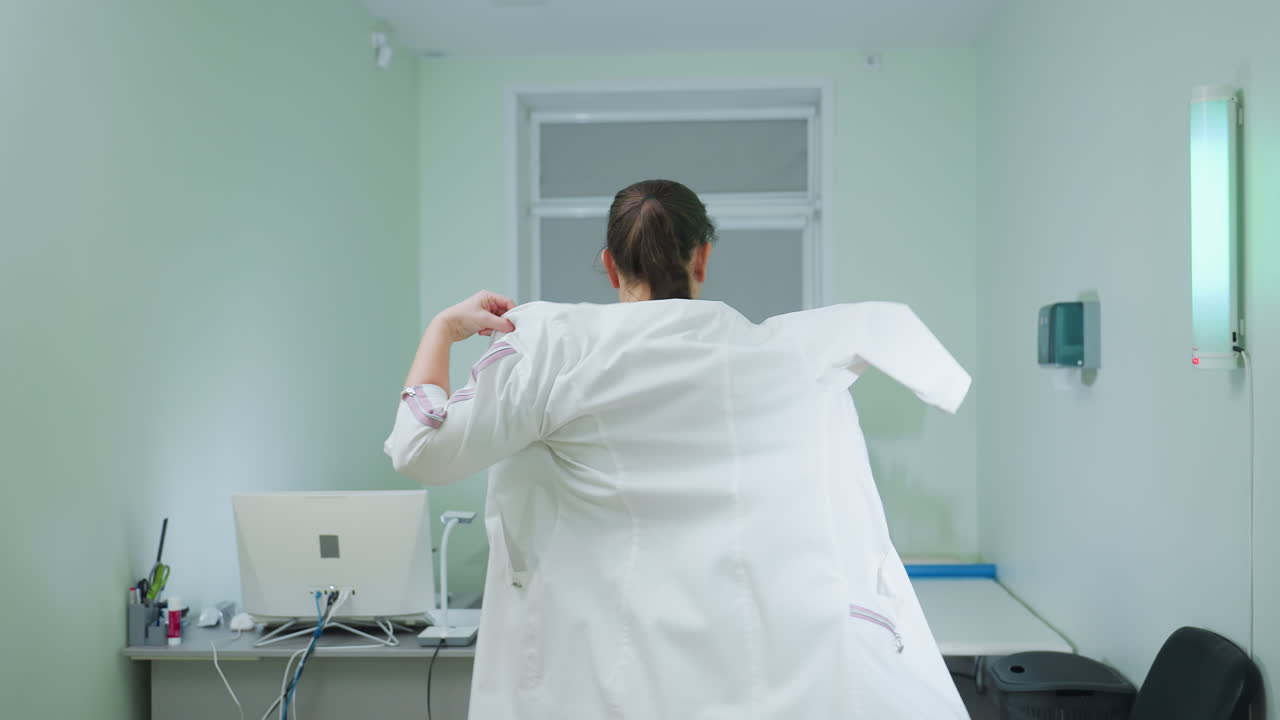 Female healthcare professional puts on uniform and walks confidently toward seat in front of desktop computer in brightly lit medical room equipped with examination table and ultrasound equipment