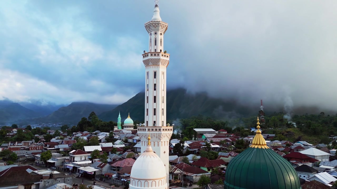Masjid Islam dengan kubah hijau dan menara menjulang dikelilingi rumah-rumah desa di bawah pegunungan vulkanik yang tertutup awan, rekaman drone udara di Indonesia