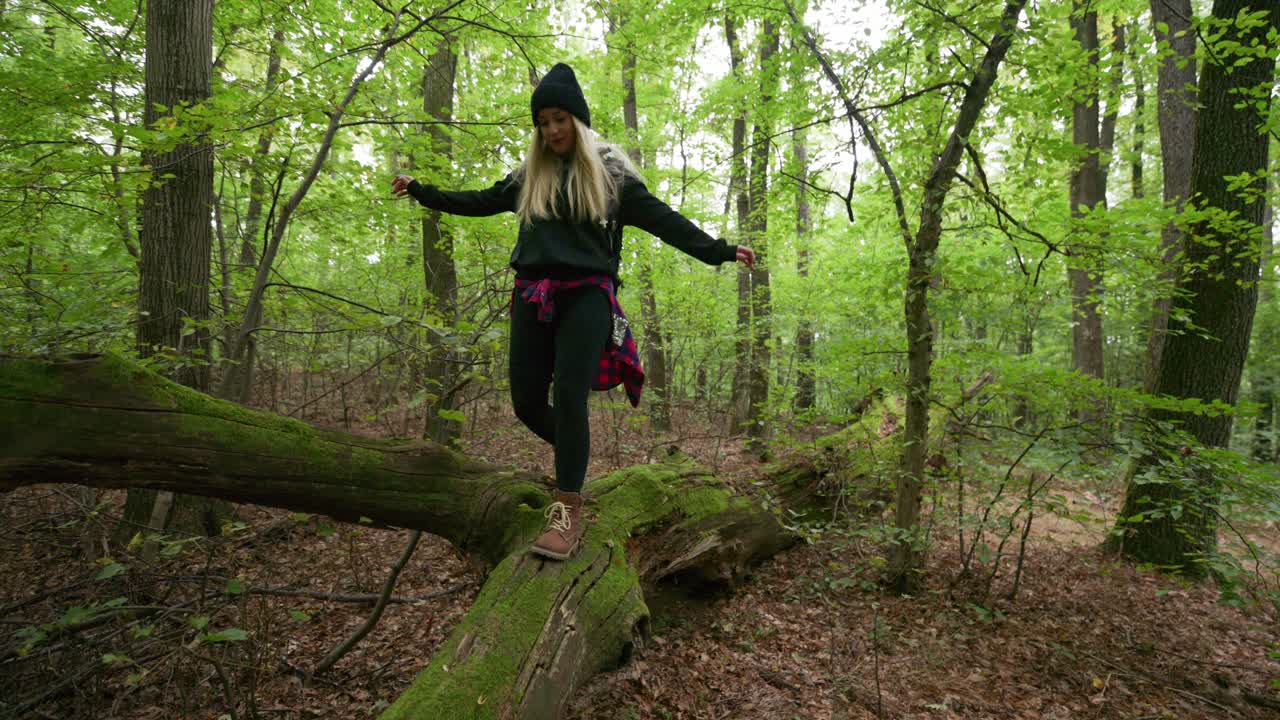 A young woman with long blonde hair wearing a beanie and casual outdoor clothing balances on a moss-covered fallen tree in the middle of a green forest