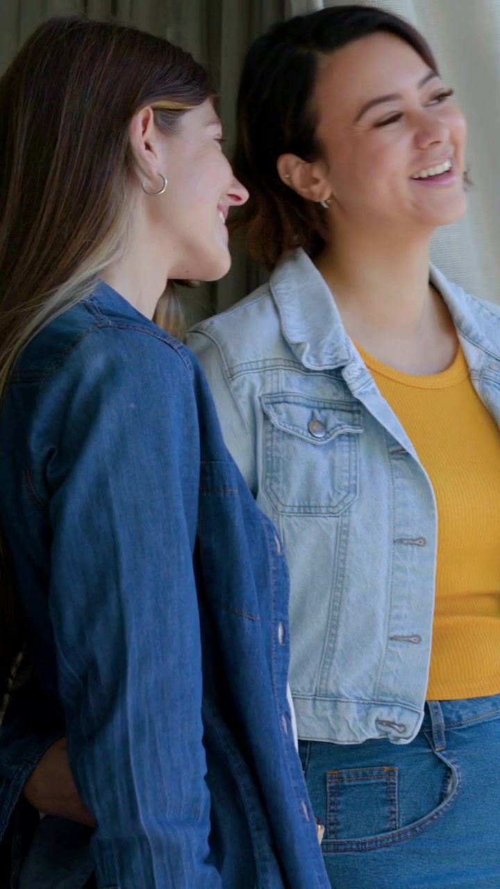 Video verticale: Due donne in denim si appoggiano e si abbracciano vicino a una porta scorrevole in vetro, l'inquadratura si stringe.