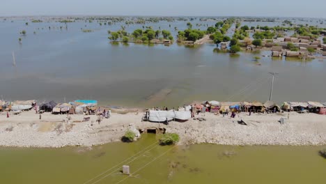 Vista-Aérea-De-La-Carretera-Elevada-Solitaria-Que-Alberga-Campamentos-Improvisados-Para-Refugiados-De-Inundaciones-Rodeados-Por-Aguas-De-Inundación-Expansivas-En-La-Zona-Rural-De-Jacobabad,-Sindh