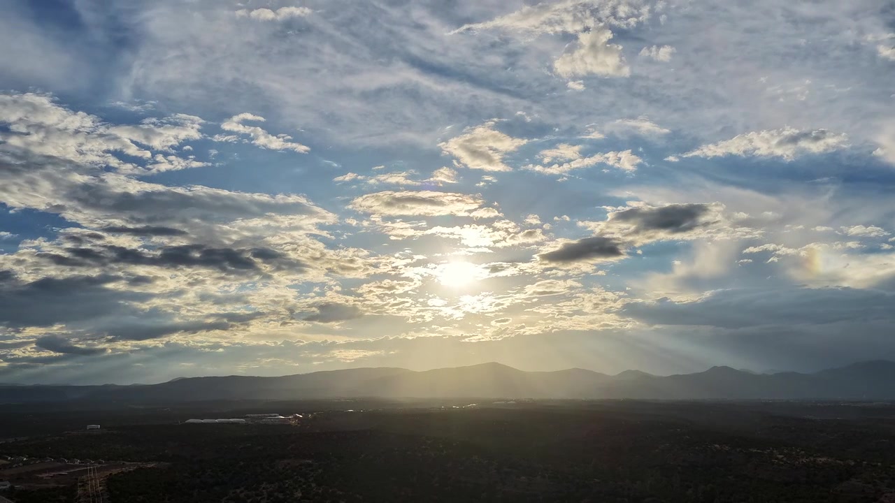 Timelapse 4K con drone di un sole sereno e luminoso che si fa strada attraverso più strati di nuvole sparse. Morbidi raggi di sole illuminano la silhouette velata delle montagne Jemez attraverso la contea di Los Alamos