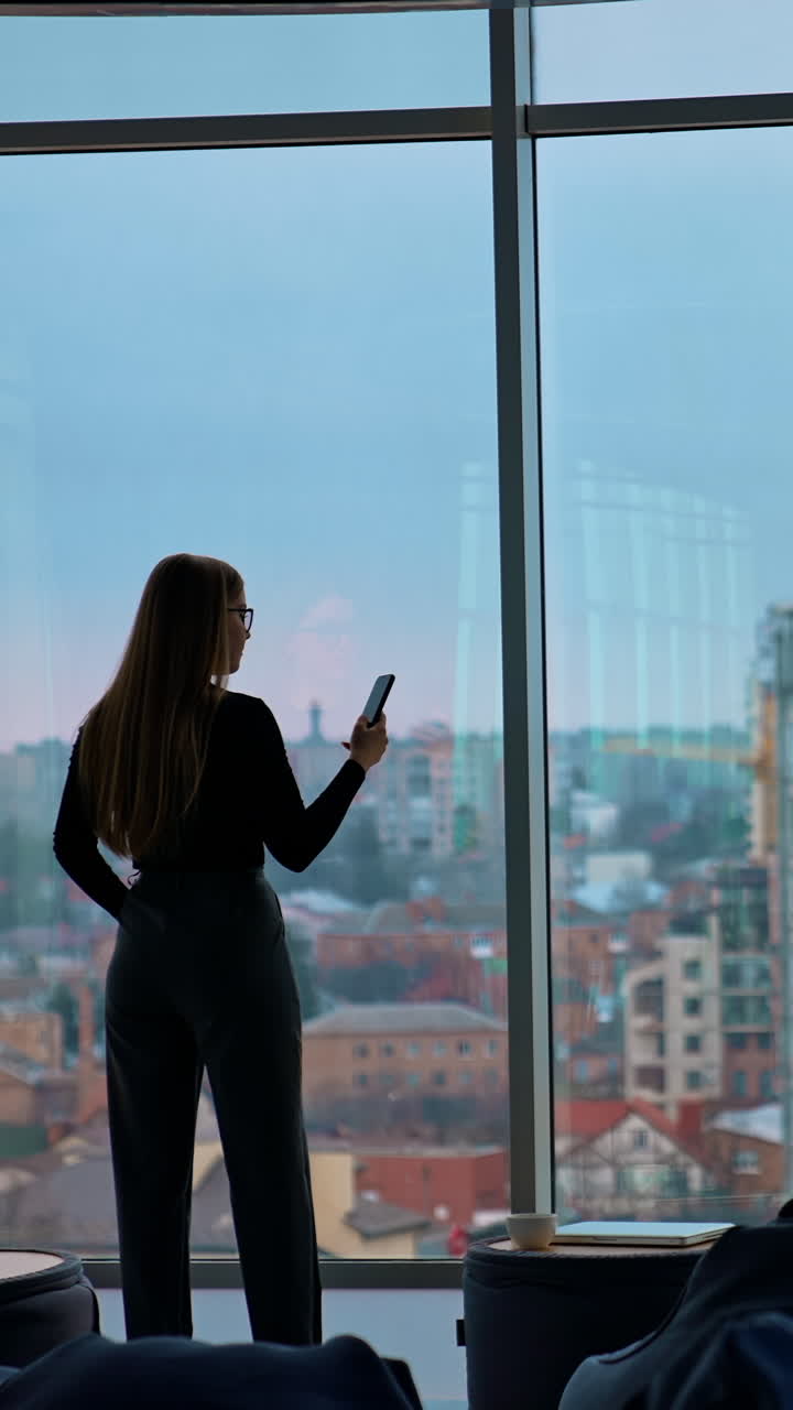 Young business woman with a phone. Rear view of a female in modern office near the large windows. Panoramic windows view with a city background. Back view. Vertical video
