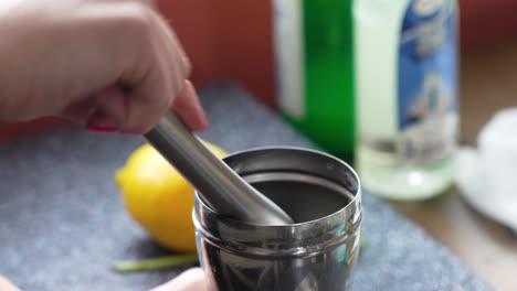 bartender twists and muddles, presses and twists in a silver cobbler shaker with a muddler to make a cocktail or mixed drink, closeup of beverage and woman’s hands tapping the edge of the glass