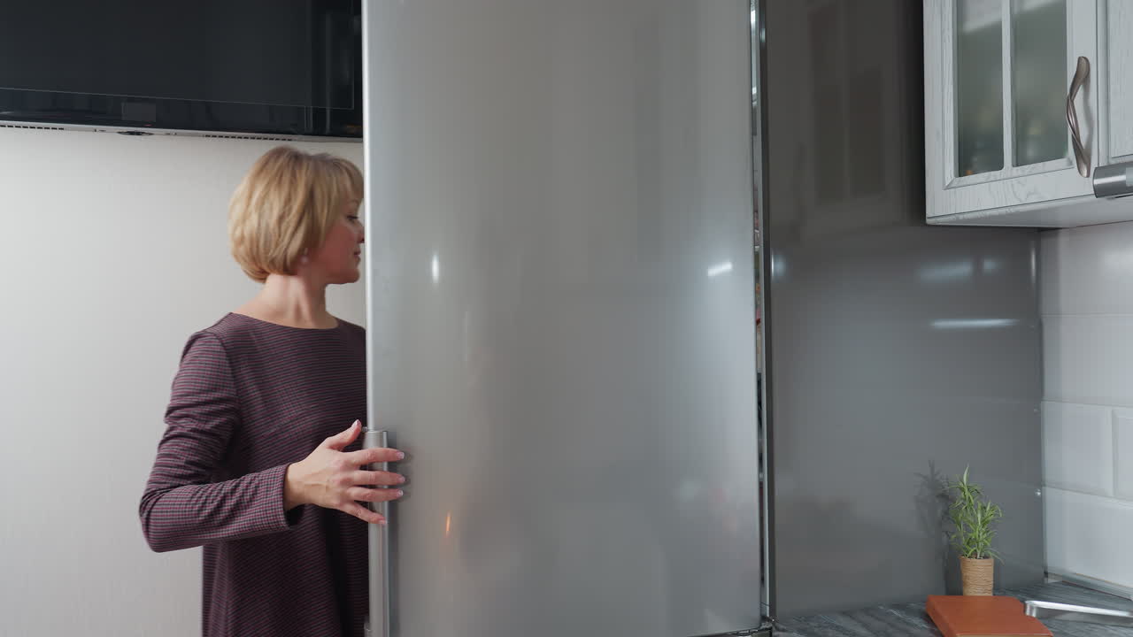 mujer joven abre la nevera, su reflejo visible en la puerta, luego la cierra mientras sostiene la caja de huevos y la coloca en la mesa, cocina moderna con electrodomésticos elegantes, decoración mínima y iluminación suave