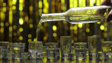 barman pour frozen vodka from bottle into shot glass. ice cubes against shiny gold party background