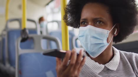 mujer de negocios afroamericana con máscara facial hablando en un teléfono inteligente y sentada en el autobús