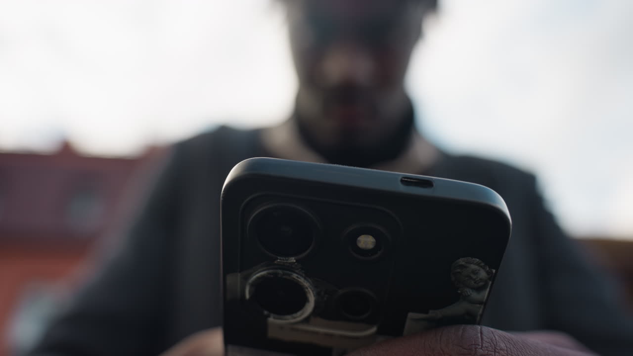 Clear view of triple camera lens on smartphone held by blurred adult outside street, showcasing modern lens detail, and sleek technology symbolizing connectivity, innovation, and digital lifestyle