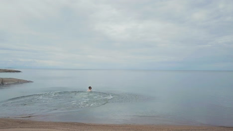 Tracking-Aerial-Man-runs-on-beautiful-ocean-beach-and-dives-into-calm-water