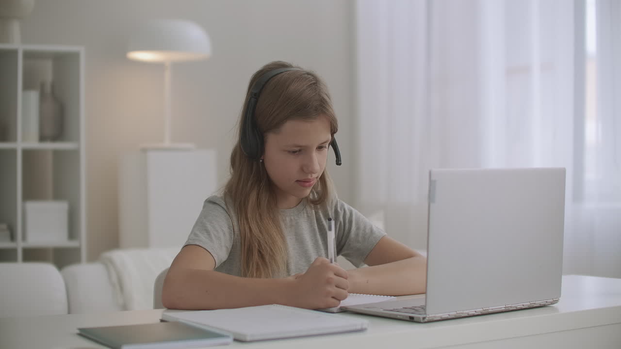 homeschooling para niños en línea chica se está comunicando con el maestro por auriculares y video chat en el portátil escribiendo en el cuaderno