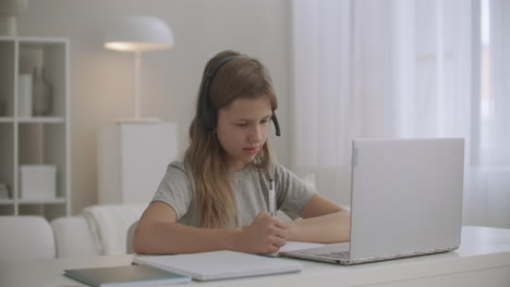 homeschooling for children online girl is communicating with teacher by headphones and video chat on laptop writing in copybook