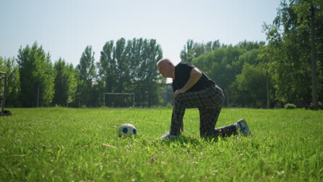 an elderly man kneels on one knee in a lush green field, focusing on fixing his shoe while a soccer ball lies close by with trees in the background