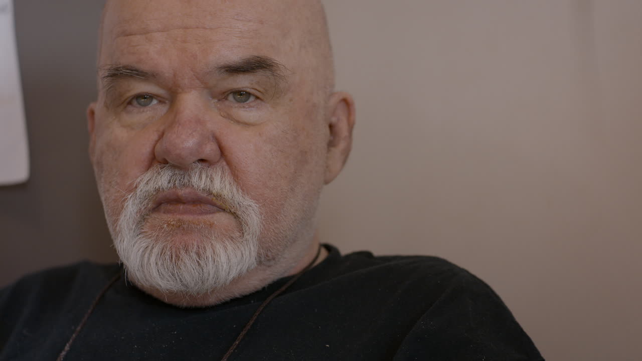 A senior male wearing a black shirt, sitting in a corner of a kitchen nook. He looks around before giving a big yawn out of boredom. He is bald, fat, with well-trimmed goatee.