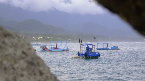 asian fishing boats bobbing on gentle ocean waves with mountains in backgrounds and rocks in foreground, filmed in handheld style as medium shot