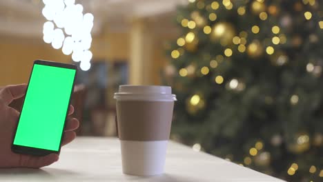close-up de la mano de un hombre sosteniendo pantalla verde smartphone maqueta. cerca de la taza de papel café para ir. fondo árbol de navidad. hombre con teléfono. llave de croma teléfono celular pantalla verde nuevo concepto de tecnología.