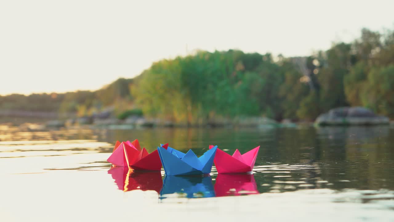 several multi-colored origami in the shape of ships are floating on the water in the lake in the evening on the background of the summer landscape