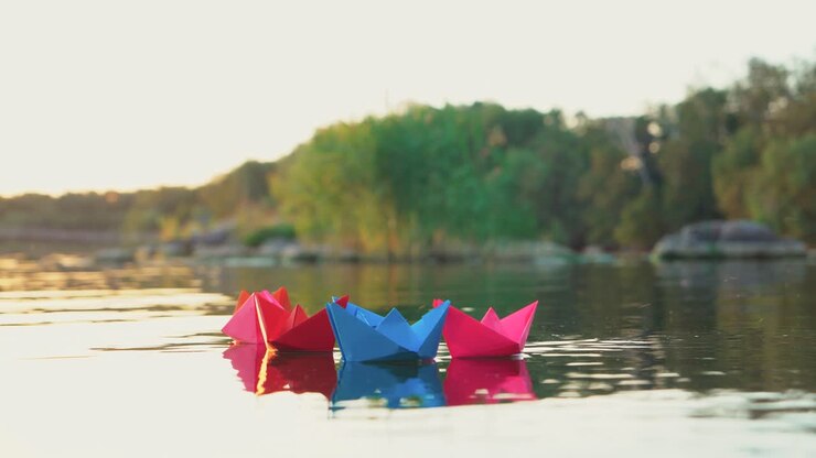 several multi-colored origami in the shape of ships are floating on the water in the lake in the evening on the background of the summer landscape