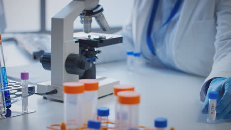 Close-Up-Of-Lab-Worker-Conducting-Research-Using-Microscope-Holding-Test-Tube-Labelled-Delta