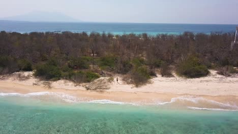 Hombre-Solitario-En-La-Playa-De-Arena-De-Una-Exótica-Isla-Deshabitada,-Retira-La-Antena-Revelando-Toda-La-Costa,-Agua-De-Mar-Turquesa-Con-Arrecifes-De-Coral-Y-Horizonte