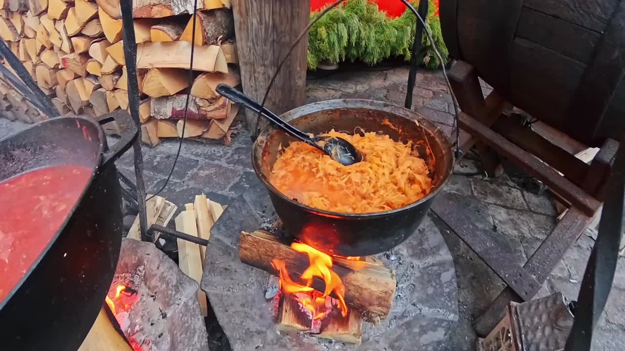 Açık hava pazarında, kepçe ile karıştırılan lahana güveci dökme demir kazanda açık ateşte kaynıyor, etrafında yığılmış odunlar ve duman var