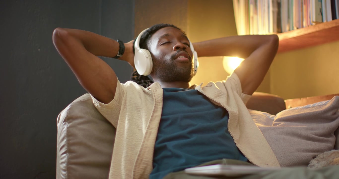 African American adult man closing laptop after working, wearing headphones, stretching on sofa