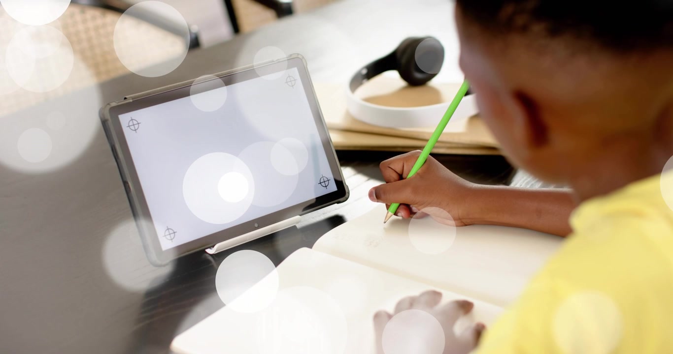 Boy sitting at desk glancing at blank tablet then writing notes in notebook for study