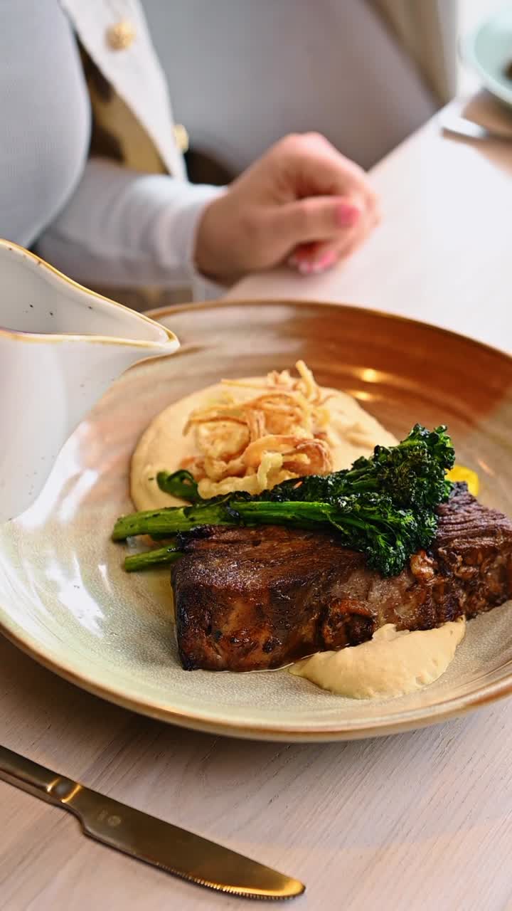 Pouring dark sauce over braised beef short rib. Motion shot of brown gravy being poured from white pitcher onto meat with vegetables